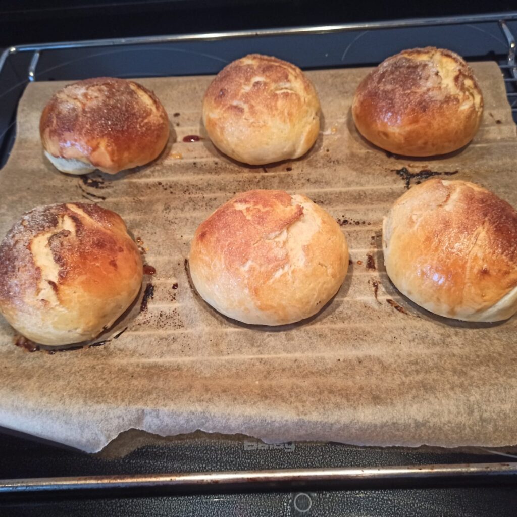 Bambas de roscón de Reyes recién horneadas, doradas y esponjosas sobre bandeja Bambas de masa estilo roscón de Reyes ya horneadas, con un acabado dorado y ligeramente craquelado, colocadas sobre una bandeja con papel vegetal. Tras el horneado, estos bollos adquieren una textura esponjosa y un aroma característico, listos para enfriar y rellenar de nata montada.